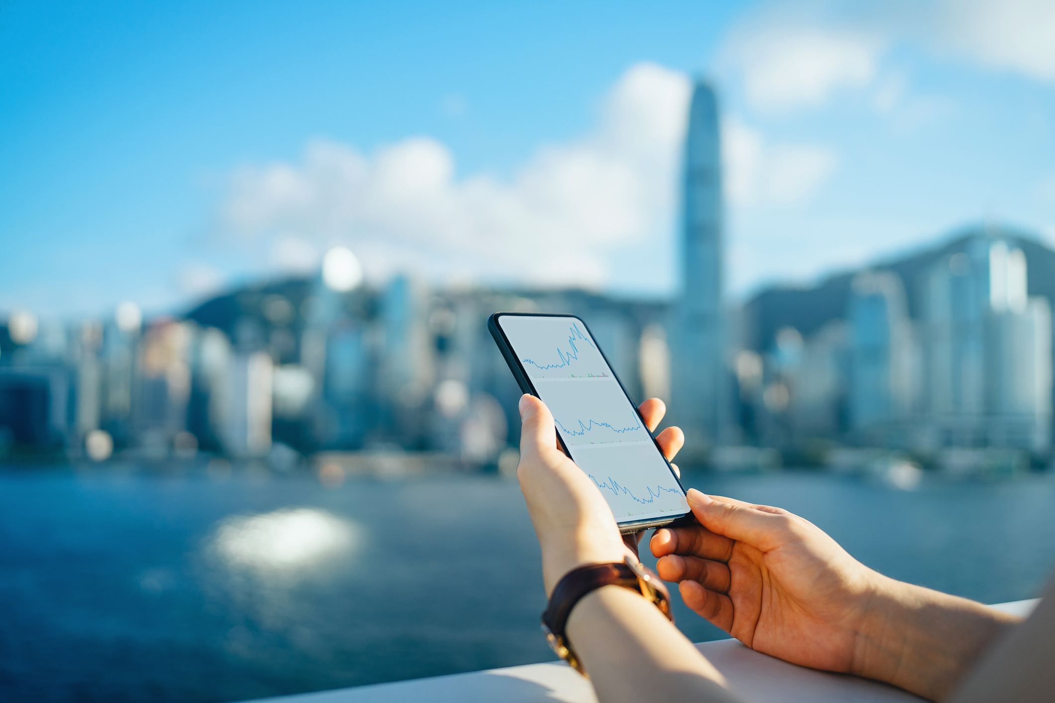 Close Up Of Woman Reading Financial Trading Data On Smartphone Against The Iconic City Skyline Of Hong Kong By The Promenade Of Victoria Harbour On A Sunny Day